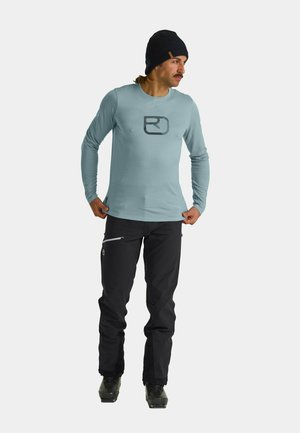 Himmelblaues Langarmshirt mit schwarzem Logo, kombiniert mit schwarzen Hosen mit einer Reißverschlusstasche. Modell trägt schwarze Stiefel und eine Mütze.