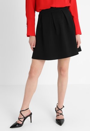 Zwarte geplooide rok van gladde stof, met een nauwsluitende tailleband. Gecombineerd met een rode blouse en zwarte hoge hakken.