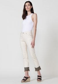 Jeans color crema con adornos de cuentas decorativas en los puños, combinados con una blusa blanca sin mangas ajustada y sandalias de cuña negras.