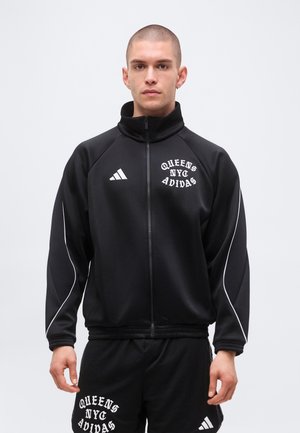 Lühikeste juustega mees kannab musta Adidas spordijopet ja lühikesi pükse, millel on kiri „Queens NYC Adidas“, seisab ühetoonilise heleda tausta ees.