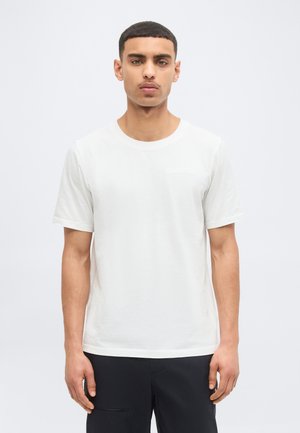 Homme aux cheveux courts portant un T-shirt blanc uni à manches courtes et un pantalon noir, se tenant devant un arrière-plan gris clair.