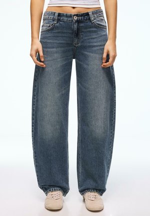 Személy laza, kék farmernadrágot, fehér tornacipőt és rövidített fehér felsőt visel, karjai lazán az oldalán pihennek.