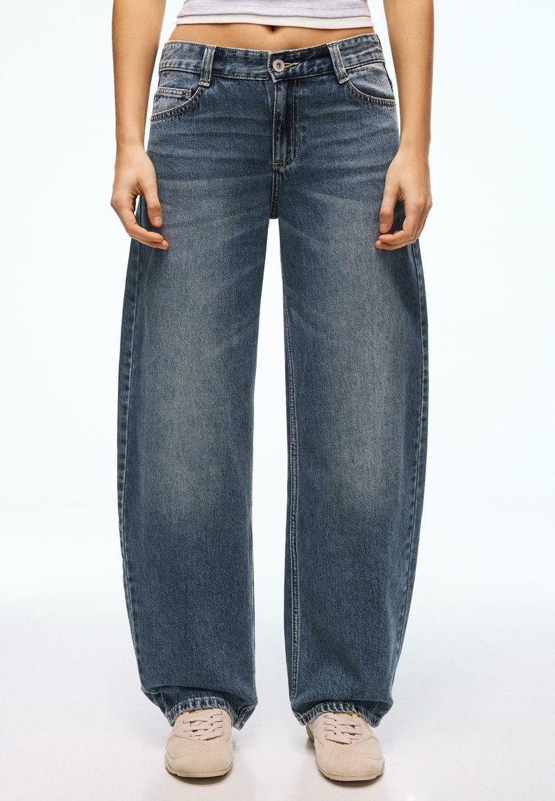 Personne portant un jean denim bleu ample, des baskets blanches et un haut blanc court, debout avec les bras détendus le long du corps.