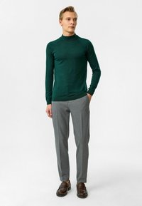 Grüner Langarm-Pullover mit hohem Kragen, getragen zu grauer Hose. Der Pullover hat eine taillierte Passform und eine gerippte Struktur. Braune Loafers runden das Outfit ab.
