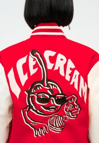 Άτομο που φοράει κόκκινο και άσπρο μπουφάν με το κείμενο "ICE CREAM" και ένα καρτούν κεράσι με γυαλιά ηλίου που κρατάει ένα κουτάλι και μια μπάλα παγωτού.
