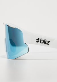 Lunettes de sport blanches avec de grandes lentilles teintées de bleu, au design élégant, et un logo noir sur le côté. Caractéristiques d'une monture durable.