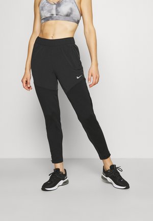 Kobieta ubrana w czarne spodnie dresowe Nike, czarno-białe buty Nike oraz szary sportowy stanik, stojąca na jednolitym tle.