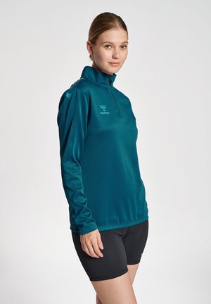 Teal langærmet pullover med lynlåse mock neck og logo på brystet. Fremstillet af glat, åndbart stof. Matchet med sorte shorts.