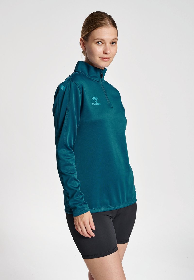 Pullover a maniche lunghe color teal con collo a zip e logo sul petto. Realizzato in tessuto morbido e traspirante. Abbinato a pantaloni corti neri.