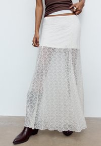 Maxinederdel i blonder i off-white med en tætsiddende ensfarvet linning, gennemsigtig tekstur, blomsterprint og en flydende silhuet, kombineret med mørkebrune støvler.