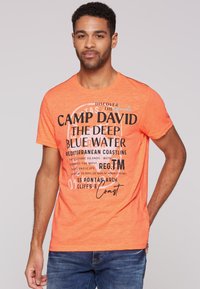 Camiseta naranja de manga corta con texto negro en negrita que presenta diversas frases, escote redondeado y una textura suave. Combinada con jeans azules.