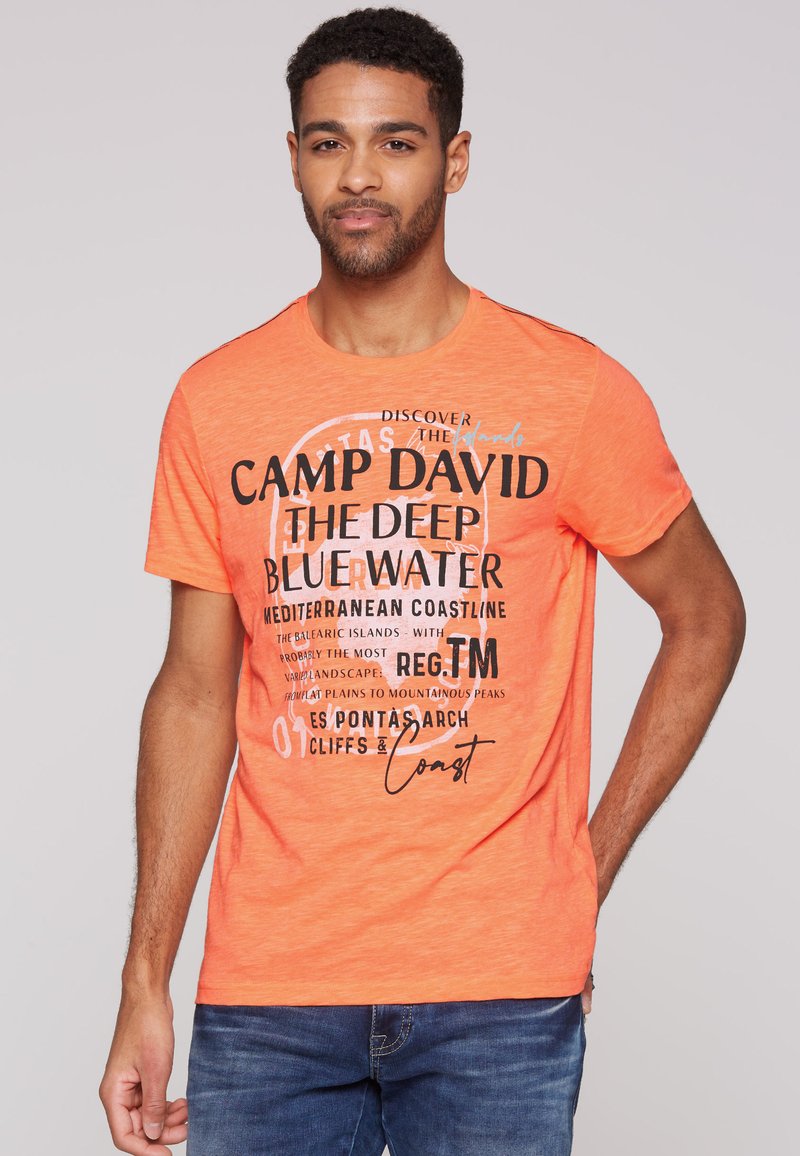 Camiseta naranja de manga corta con texto negro en negrita que presenta diversas frases, escote redondeado y una textura suave. Combinada con jeans azules.