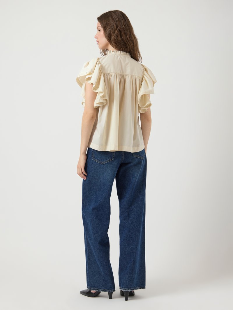 Cream blouse with ruffled short sleeves, gathered back, paired with wide-leg blue jeans and black heels, showcasing a relaxed fit.