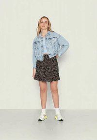 Casaco de denim azul claro com dois bolsos no peito, combinado com uma saia preta de flores. O modelo usa ténis brancos com detalhes amarelos.