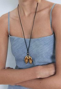 Collier pendentif doré en forme de feuilles abstraites, suspendu à un cordon noir, porté sur une robe en denim clair à fines bretelles.
