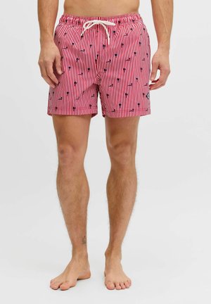 Man draagt rood-wit gestreepte zwembroek met kleine zwarte palmboom patronen en een wit trekkoord, staand op blote voeten.