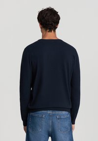 Maglione blu navy realizzato in un fine tessuto a maglia, con scollo rotondo e maniche lunghe. Texture liscia con una vestibilità classica. Indossato sopra jeans blu.