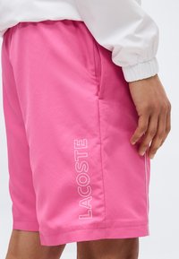 Άνδρας που φοράει φωτεινά ροζ σορτς με την επιγραφή «LACOSTE» κάθετα κοντά στην αριστερή τσέπη, σε συνδυασμό με λευκό μακρυμάνικο μπλουζάκι.