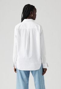 Chemise blanche à manches longues avec un col classique et un ourlet arrondi, présentant une couture de yoke à l'arrière et des poignets à bouton.