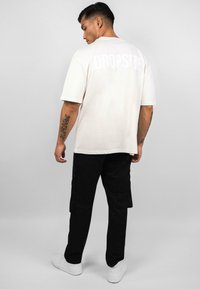 Weißes Baumwoll-T-Shirt mit "DROPSIZE" auf dem Rücken, kombiniert mit schwarzen Cargo-Hosen und weißen Sneakers. Modell ist von der Kamera abgewandt.