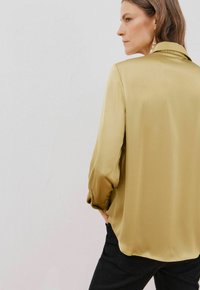 Camicia a maniche lunghe in seta di colore verde oliva, con colletto e maniche con polsini. Indossata con pantaloni neri, texture liscia visibile.