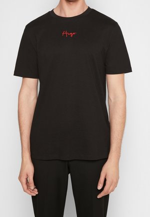 Homme portant un t-shirt noir uni à manches courtes avec un petit logo rouge "Hugo" brodé au centre de la poitrine.