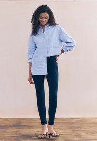 Světle modrá a bílá pruhovaná košile s knoflíky a kapsou, kombinovaná s tmavě modrými slim-fit kalhotami a černými žabkami.