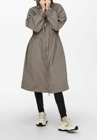 Person i en lång, lös grå kappa med dragkedja fram, svarta byxor och vita chunky sneakers, stående mot en neutral bakgrund.