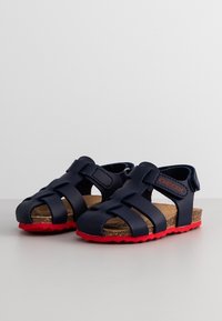 Lasten tummansinisiä sandaaleja punaisilla pohjilla, korkkitutkilla ja säädettävillä tarranauhoilla, sijoitettu vierekkäin valkoiselle pinnalle.