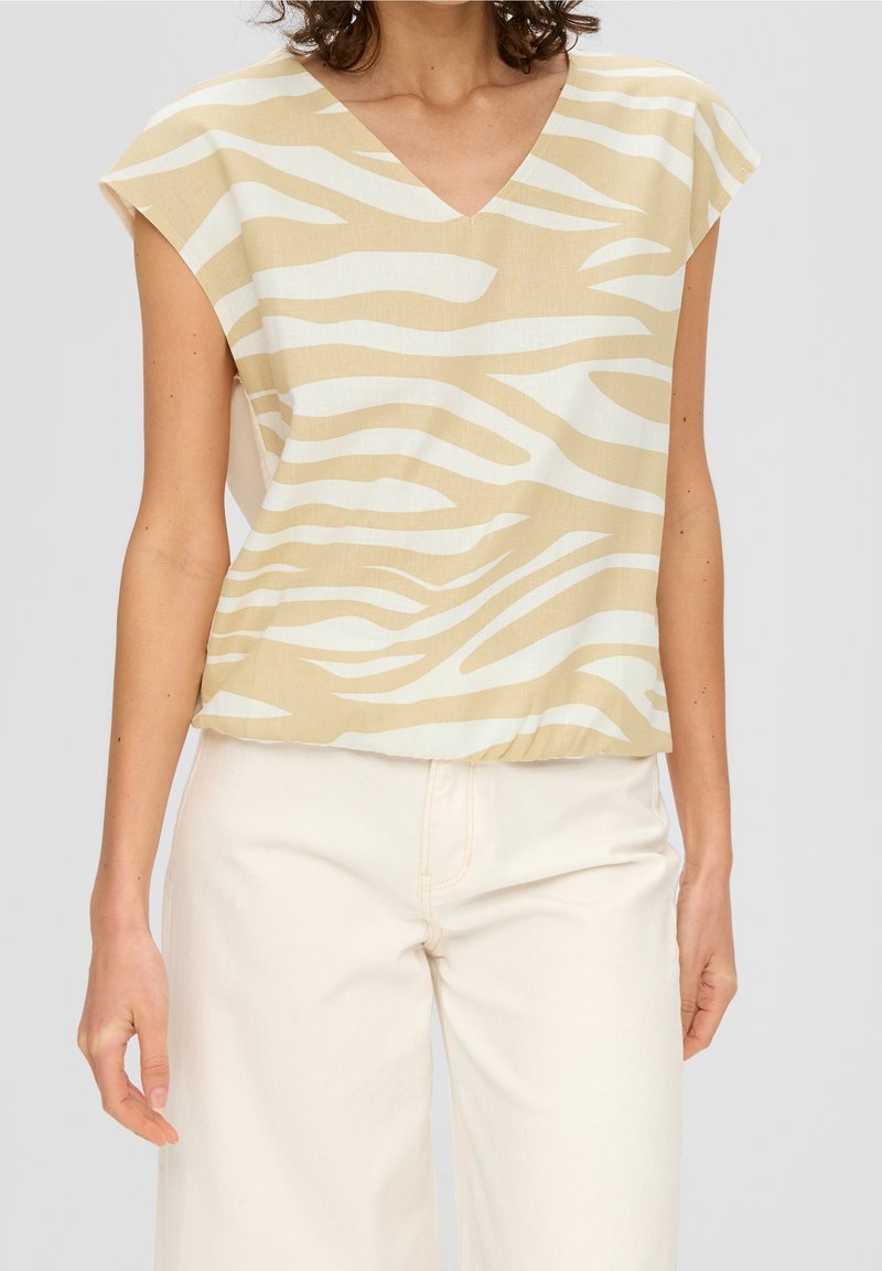 Femme portant un haut à manches courtes beige et blanc à rayures zébrées avec décolleté en V, associé à un pantalon crème taille haute sur fond gris clair.