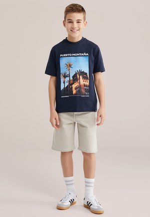 Usmievajúci sa chlapec stojí a má na sebe tmavomodré tričko s potlačou palmy a budovy, béžové šortky, biele ponožky s pruhmi a biele tenisky na neutrálom pozadí.