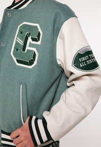 Green wool-blend varsity jacket with cream leather sleeves, featuring an embroidered "C" logo and striped ribbed cuffs and waistband.