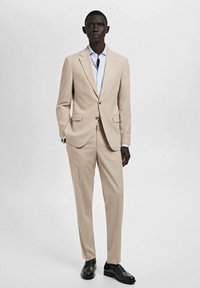 Completo beige a due pezzi con rever a puntale, chiusura con un solo bottone e pantaloni dritti. Abbinato a una camicia azzurra e scarpe nere.