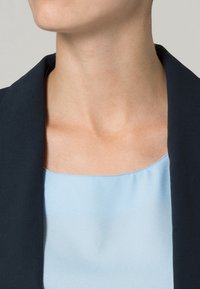 Marinblå blazer med slagkrage över en ljusblå, slät satinblus. Synlig axelsöm och fyrkantig halsringning. Hudton synlig vid halsen.