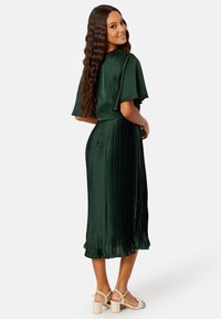 Robe en satin vert avec jupe plissée, manches flottantes et texture lisse. Détails notables comprennent une silhouette fluide et une longueur mi-mollet.
