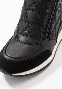 Svart sneaker i quiltat läder, små metallnitar, sidodragkedja och vit sula, visad i närbild mot en vit bakgrund.