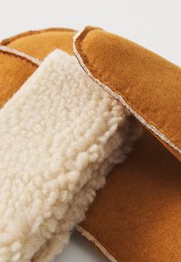 Tan wollen slippers met een textuur van beige fleece voering. Kenmerkt zich door contrasterende stiksels en een zachte, ronde vorm.