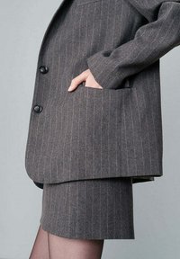 Blazer grigio a righe con due bottoni, abbinato a una minigonna grigia coordinata. Presenta una grande tasca laterale e una vestibilità su misura.