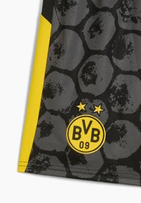Mustapohjaista ja harmaata urheiluprinttiä, jossa on teksturoitu pyöreä kuvio, kirkkaat keltaiset yksityiskohdat ja pyöreä logo, jossa lukee "BVB 09" ja kaksi tähteä.