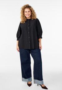 Svart blus med knappar, veckdetaljer och trekvartsärm, kombinerad med vida mörka jeans i denim med uppvikta fållar.