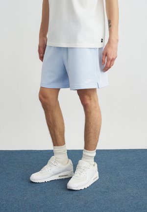 Persona que lleva pantalones cortos Nike azul claro, calcetines blancos y zapatillas Nike blancas, de pie sobre una alfombra azul.