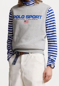 Šedá bezrukávová mikina s nápisem "POLO SPORT" modrým a červeným písmem, nošená přes modrou a bílou pruhovanou košili s dlouhým rukávem; krémové kalhoty.