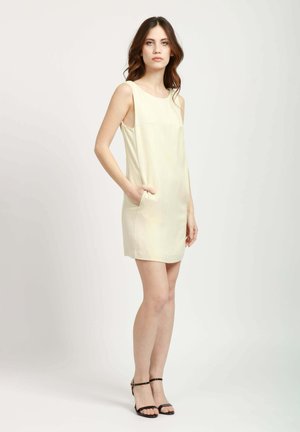 Abito senza maniche di un giallo chiaro, con una linea dritta, caratterizzato da tasche laterali e una texture morbida. Abbinato a sandali neri con cinturino alla caviglia.