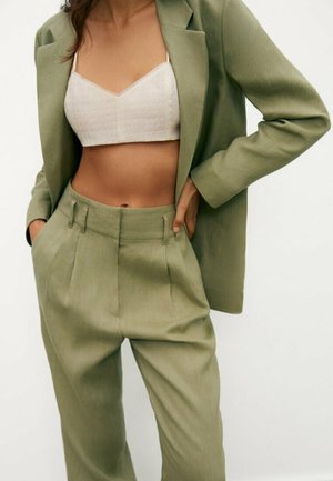 Femme portant un blazer vert clair ajusté et un pantalon plissé avec un top en maille blanc court, mains dans les poches, debout sur fond blanc.