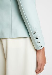 Ljusblå kavaj med skräddarsydd passform, slät tyg och fyra dekorativa silverknappar på ärmen. Vit underklädesplagg synligt.