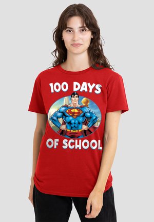 Young woman wearing a red t-shirt showing Superman and text "100 Days of School" on a plain light gray background.