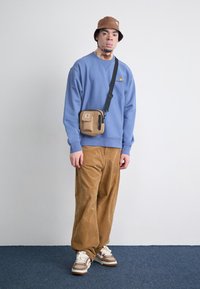 Niebieska bluza, beżowe spodnie z kordurowej tkaniny, brązowy kapelusz typu bucket i brązowa torba na ramię. Na nogach beżowe trampki z brązowymi akcentami. Prosty design.