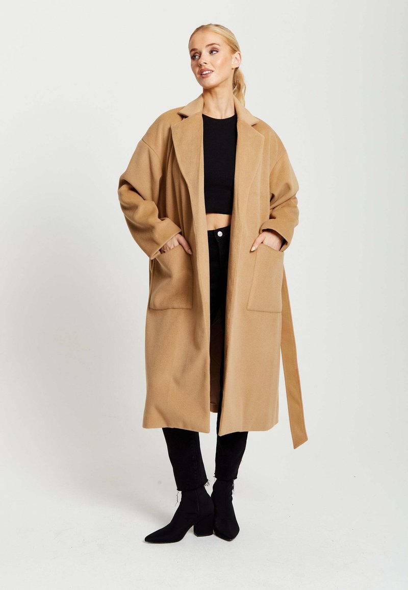 Camel-colored oversized coat with pockets, featuring a shawl collar and belted waist, paired with black cropped top and fitted black pants.