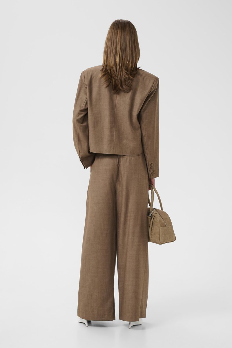 Femme aux cheveux bruns clairs et lisses, portant un costume marron surdimensionné et des chaussures blanches, tenant un petit sac à main marron, vue de dos.