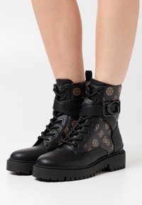 Stivali neri alla caviglia con suole spesse, motivi monogram marroni, lacci frontali e cinghie con fibbia indossati su gambe nude su uno sfondo bianco.